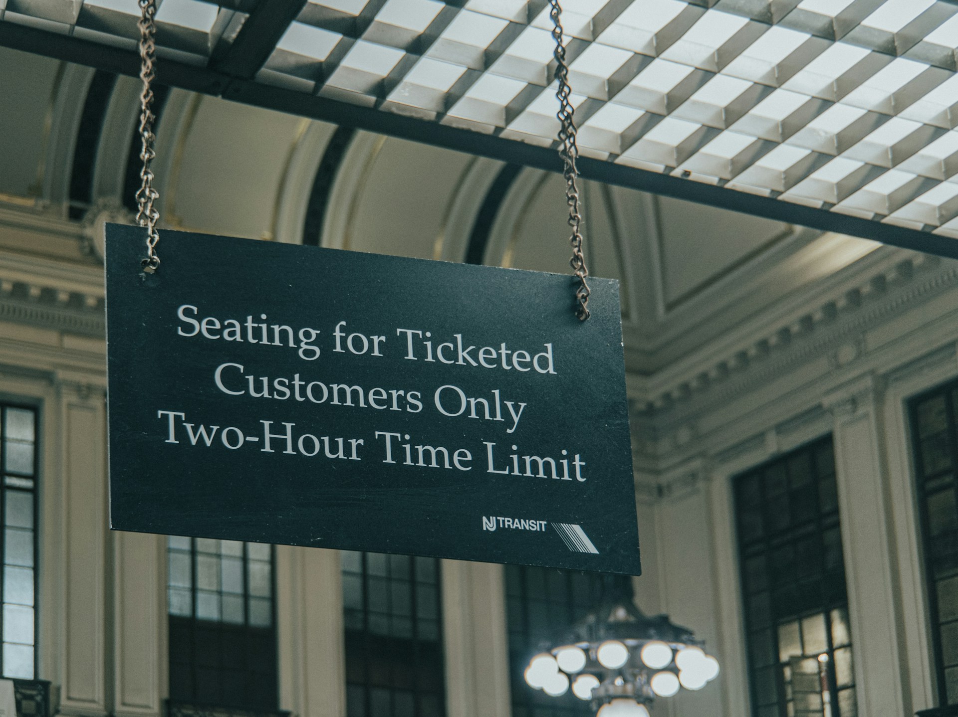 Black sign reading "Seating for Ticketed Customers Only, Two-Hour Time Limit" hangs from chains in a grand station